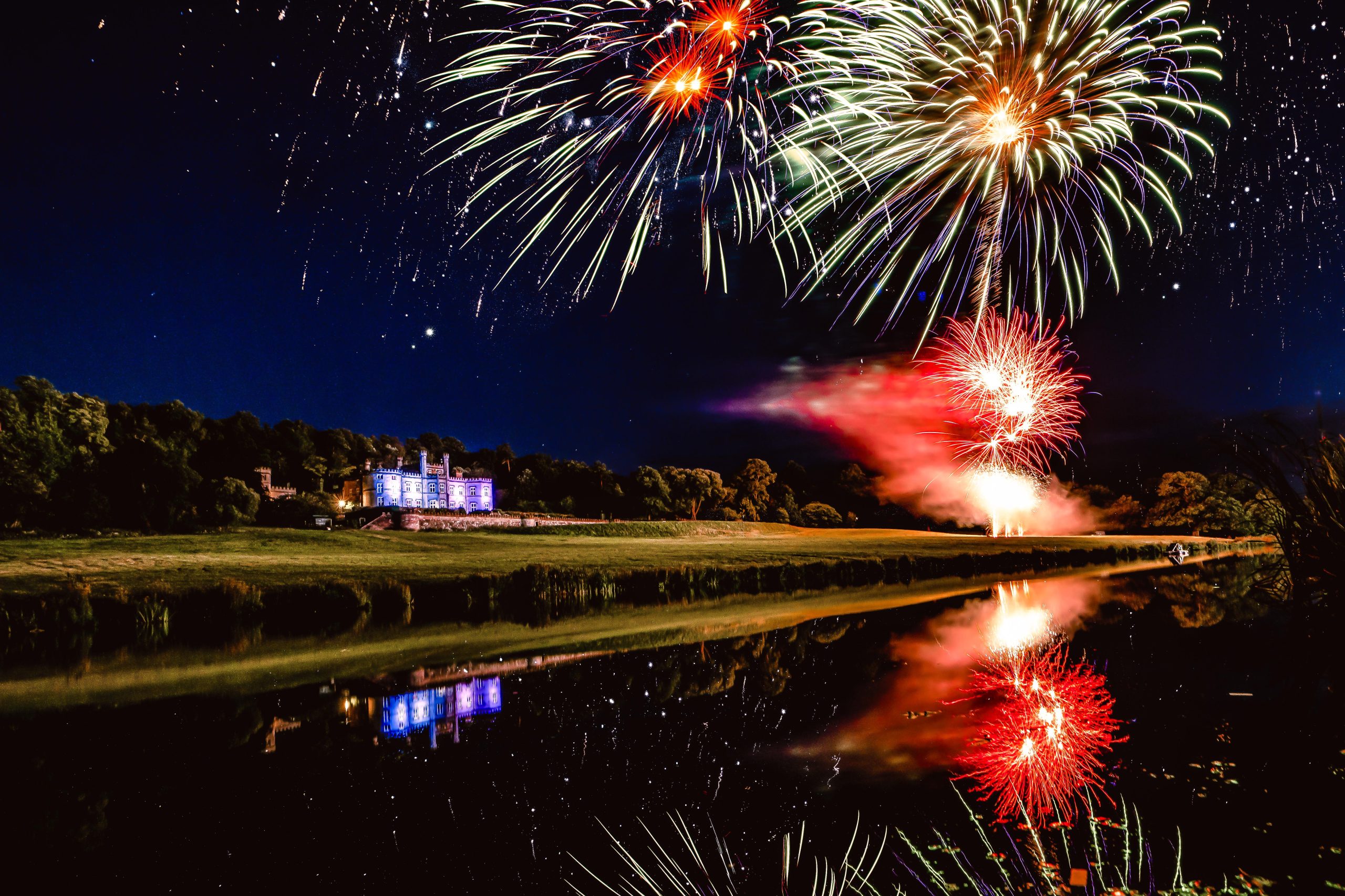 Vibrant fireworks display lighting up the night sky over Bolesworth Castle, with colourful reflections on the calm water below.