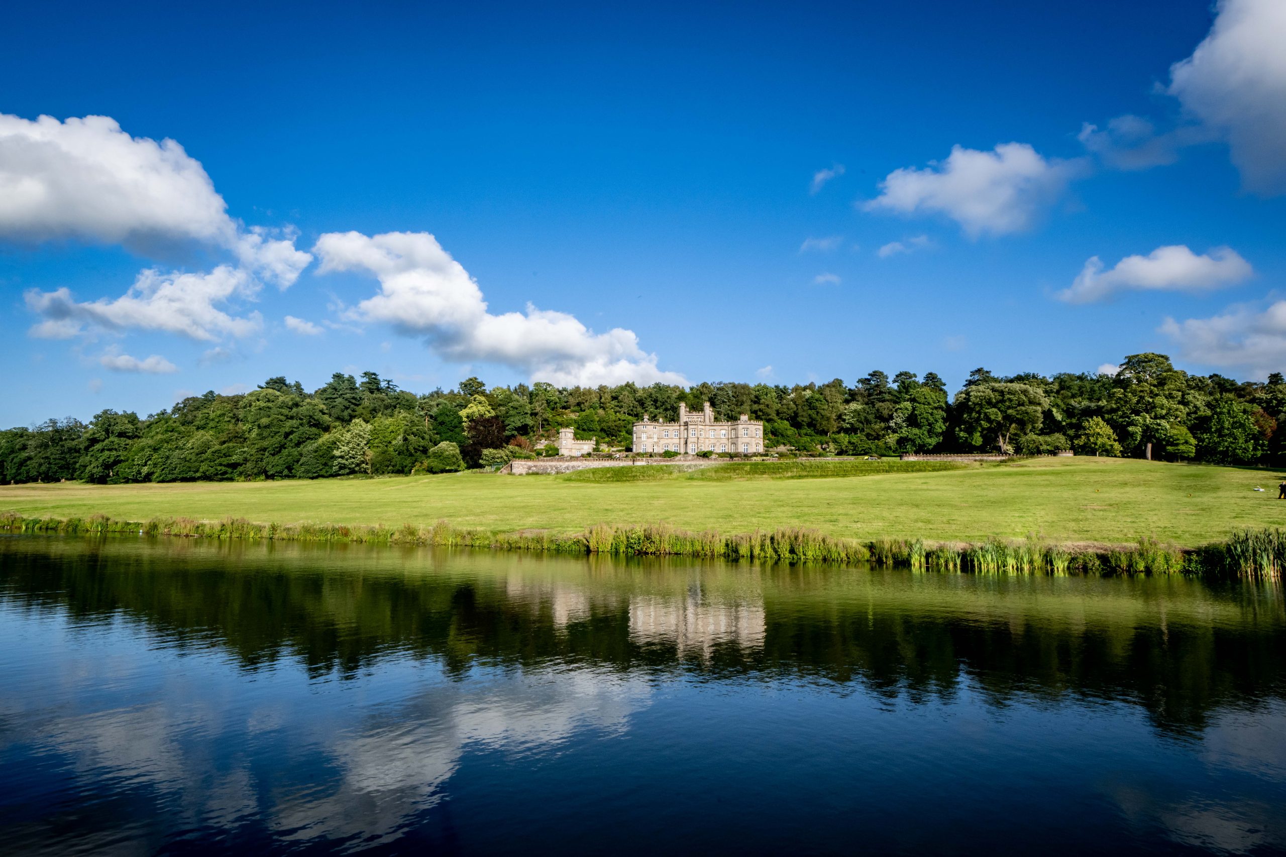 A picturesque view of Bolesworth Castle, reflected in the tranquil lake, surrounded by lush greenery and a vibrant blue sky.