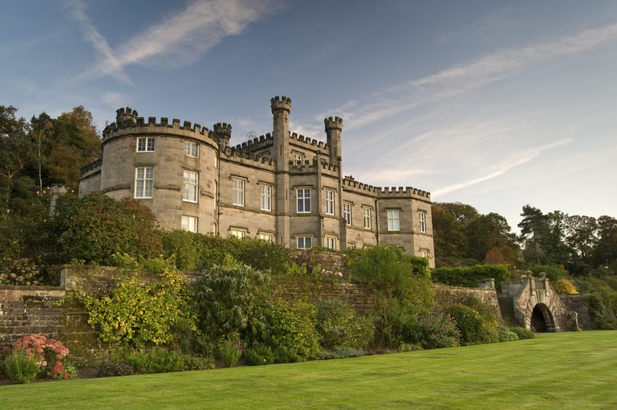 A stunning view of Bolesworth Castle, a historic estate in Cheshire, UK, surrounded by lush gardens and an expansive lawn. The castle's majestic stone architecture and picturesque landscape create an idyllic setting.