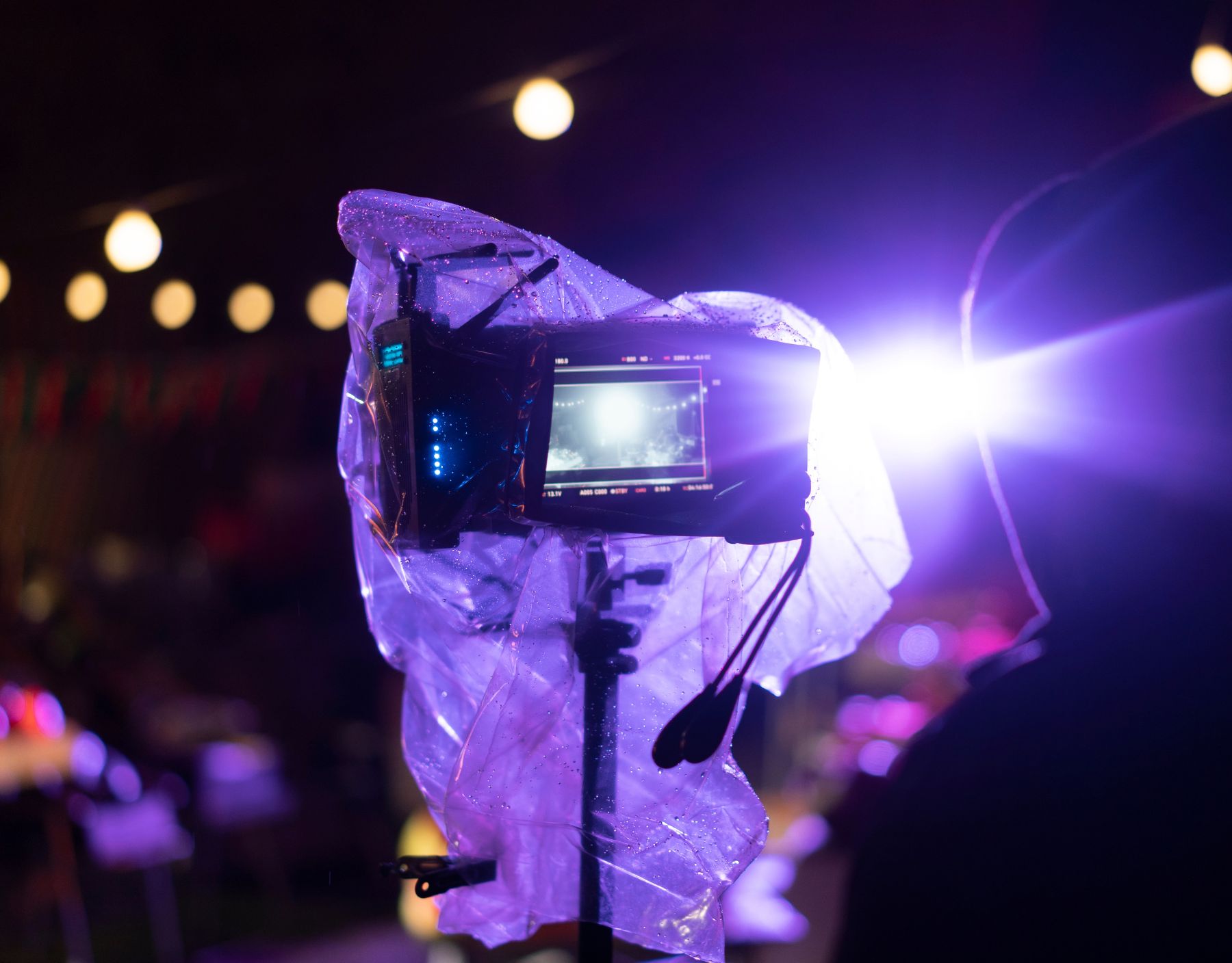 A professional camera rig covered in plastic for rain protection, capturing a cinematic scene under dramatic purple lighting. A silhouetted figure stands in the background, adding to the atmospheric behind-the-scenes feel of a film production.