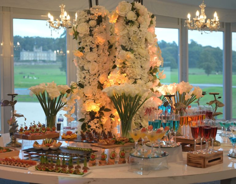 Cultural wedding setup with white flowers, elegantly arranged table settings in a marquee with chandeliers, drinks, and canapés.
