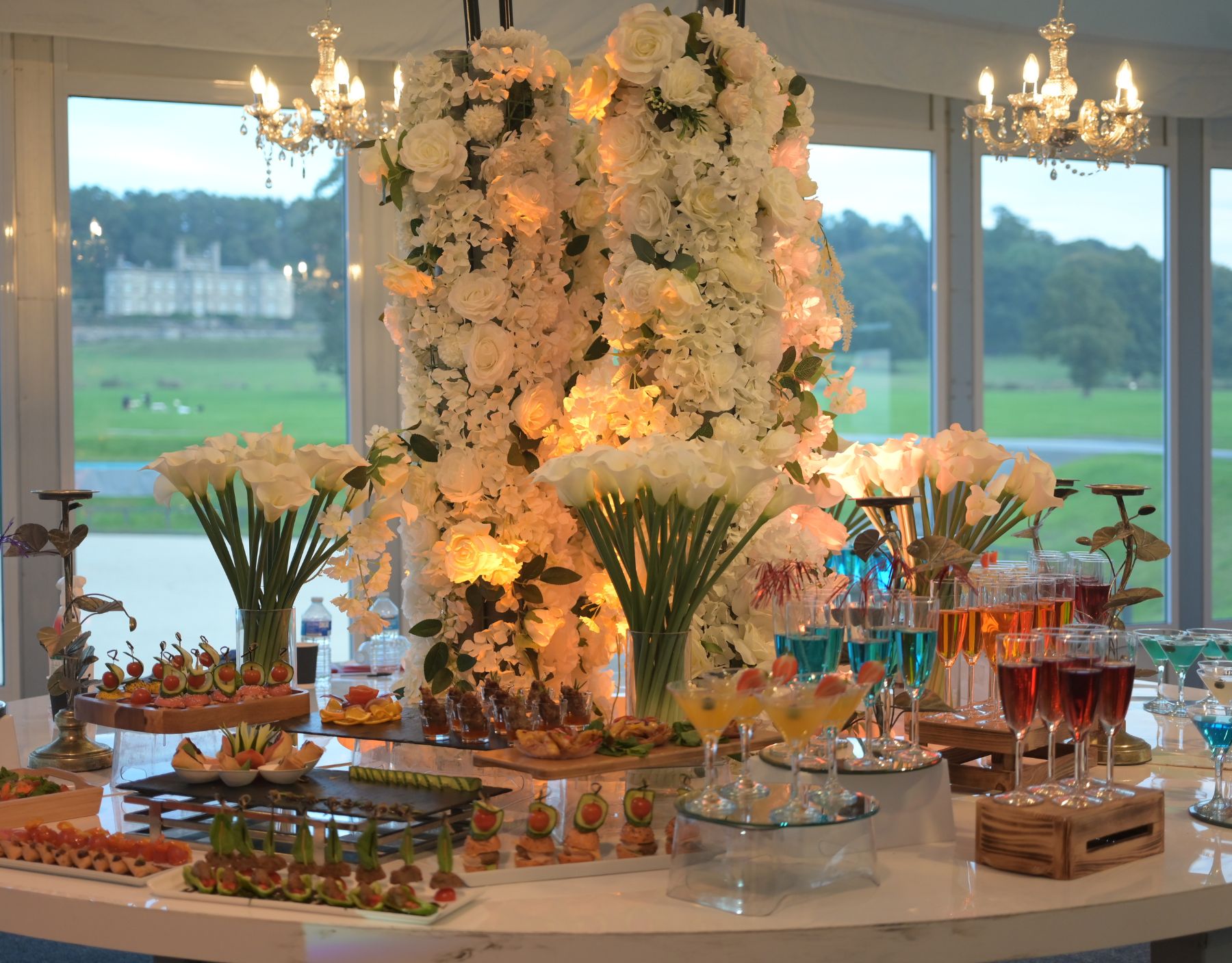 Cultural wedding setup with white flowers, elegantly arranged table settings in a marquee with chandeliers, drinks, and canapés.