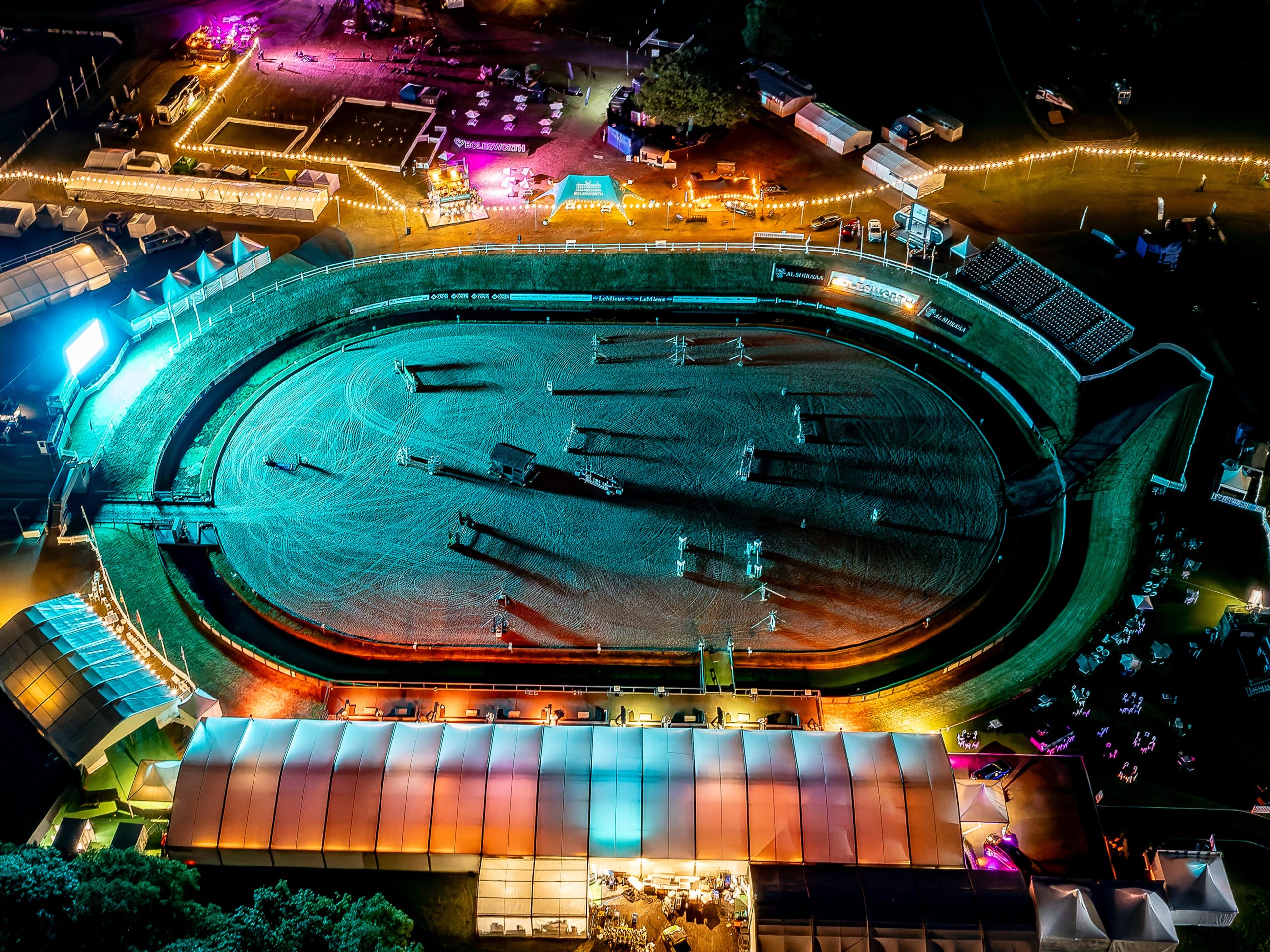 Night-time aerial view of the Bolesworth International Arena, brightly lit with colourful lights, surrounded by event structures and activities.