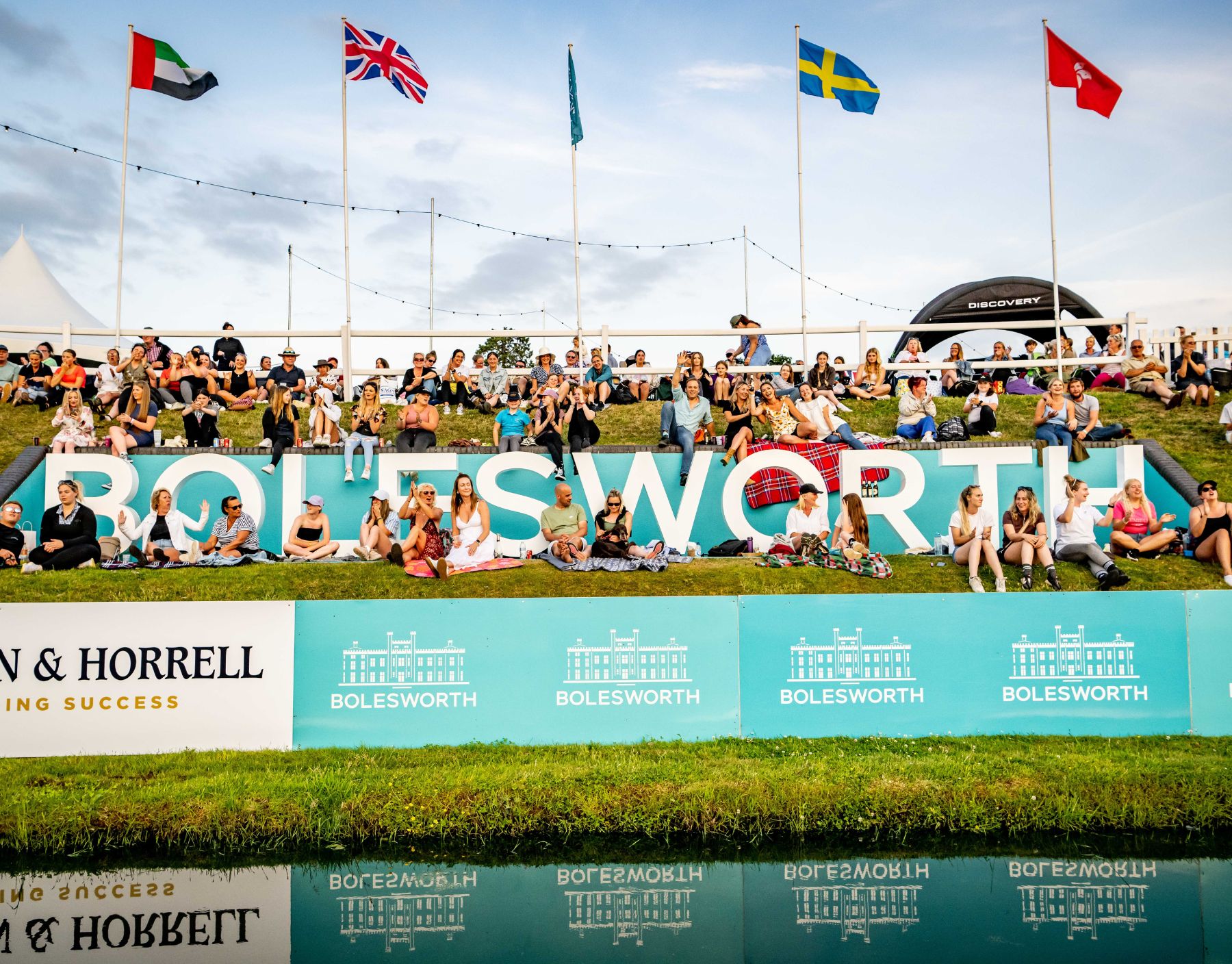 A crowd of people sitting on a bank around an equestrian show ground, cheering on riding competitions