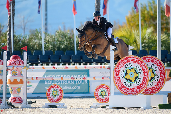 A skilled showjumper riding Harthill Magician, soaring over a colourful obstacle at a professional equestrian event. The competition arena is set against a backdrop of international flags, with a clear blue sky enhancing the high-stakes intensity of the jump.
