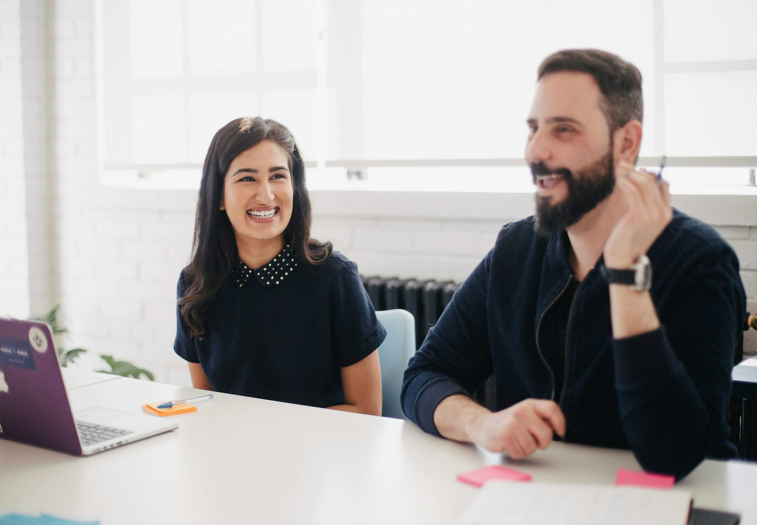 A diverse team meeting in a modern office space, with a smiling woman and a bearded man engaged in a discussion. The setting includes a bright, open workspace with a laptop, notebooks, and sticky notes, fostering a collaborative and creative work environment.