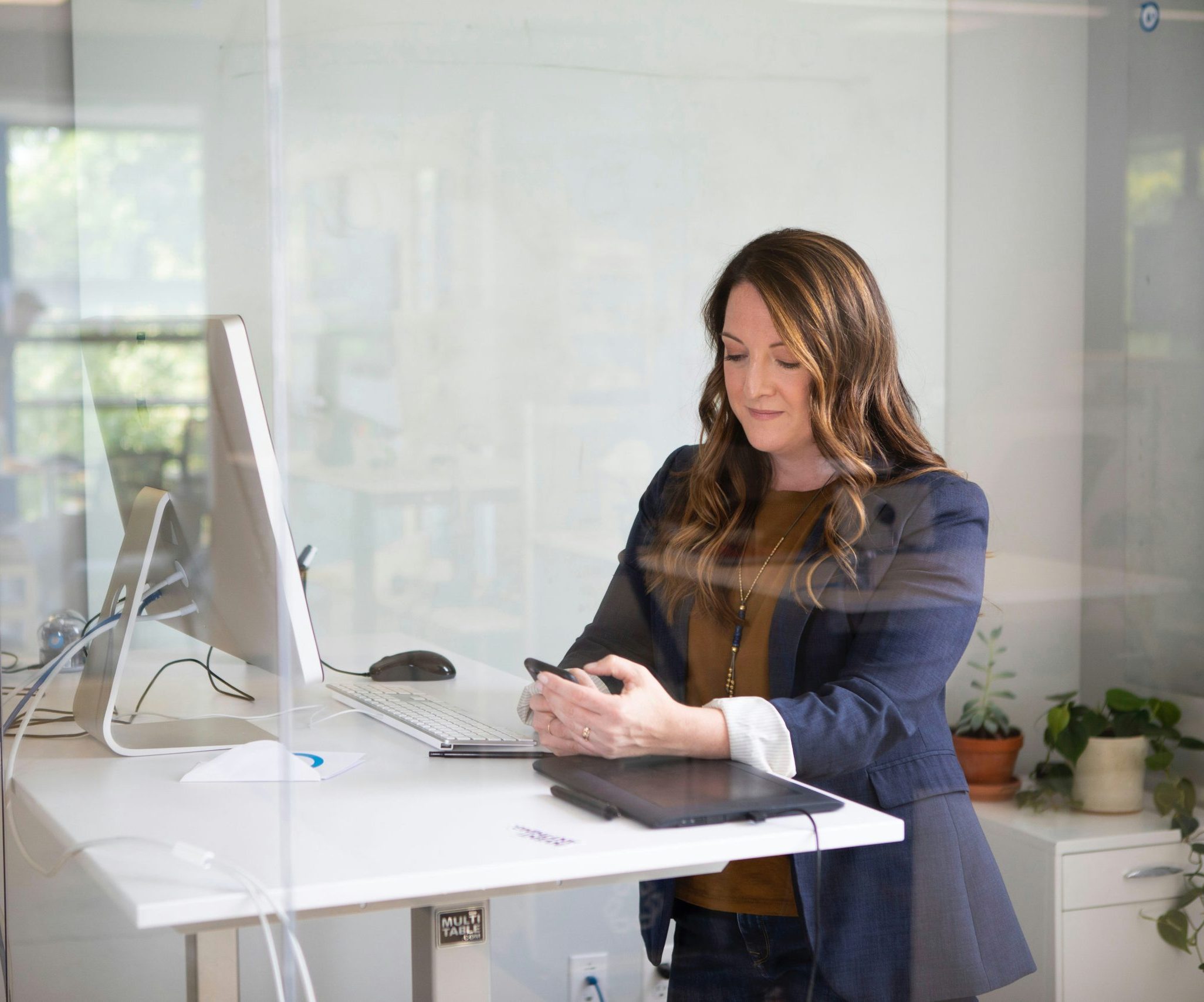 A professional woman in a modern office space, working at a standing desk while using her smartphone. The setting includes a desktop computer, tablet, and indoor plants, reflecting a productive and contemporary work environment.
