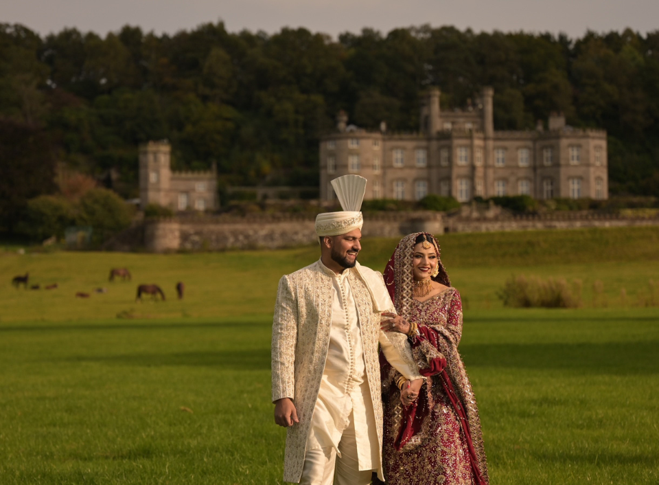 Bride and groom in traditional South Asian wedding attire walking with Bolesworth Castle in the background.
