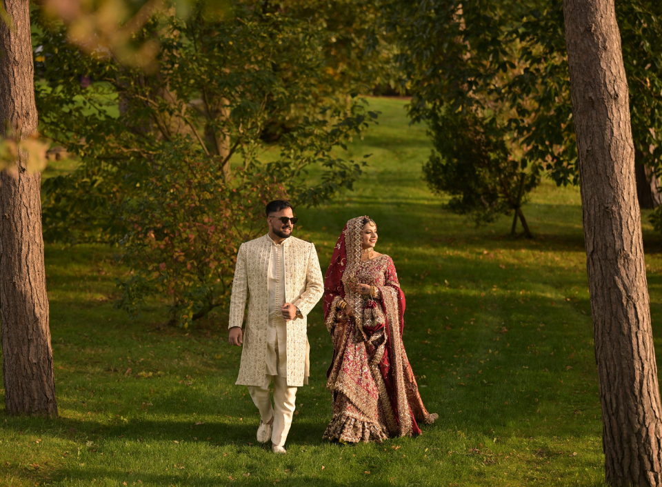 Bride and groom in traditional South Asian wedding attire walking through woodland at Bolesworth.